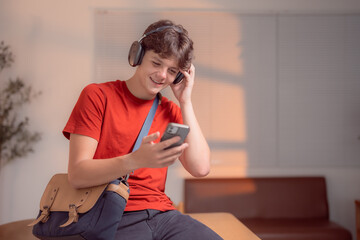 Young student enjoying music streaming with wireless headphones, selecting songs on a mobile phone app while sitting comfortably at a table in a cozy home environment
