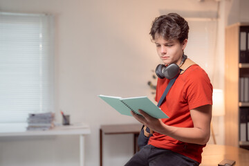 Concentrating teenager immersed in reading a book at home, headphones resting around his neck while studying at a desk, creating a cozy and focused learning environment