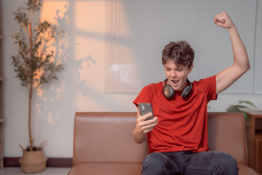 Excited teenager celebrating success on a brown sofa at home, raising a fist in joy with headphones around his neck and a smartphone in hand - Powered by Adobe