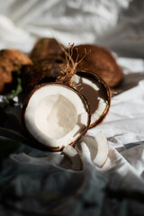On a soft white cloth, several whole coconuts lie next to a halved coconut showcasing its creamy inner flesh. Natural light highlights the textures and colors, creating an inviting scene.