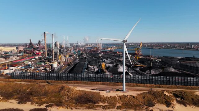 Steel production site in IJmuiden, Netherlands, featuring smokestacks, raw materials, and wind turbines. The aerial perspective highlights the tension between heavy industry and renewable energy.