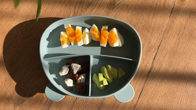 First solid food, healthy baby meal with boiled egg, avocado, bread in a silicone plate in the shape of a bear on the table. Beautiful morning sunlight and flowers nearby. 