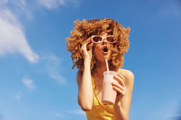 Happy woman with curly hair wearing sunglasses, enjoying a smoothie against a clear blue sky