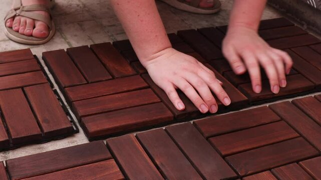 Woman connecting wooden panels on balcony floor. Installing interlocking wood tiles for outdoor upgrade. DIY balcony renovation with dark wood texture.