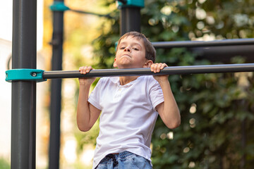 Fototapeta premium Little schoolboy, young athlete is pulling himself up on a sport horizontal bar on outdoor playground on sunny day. Active childhood, acrobat, workout on backyard, summer camp concept