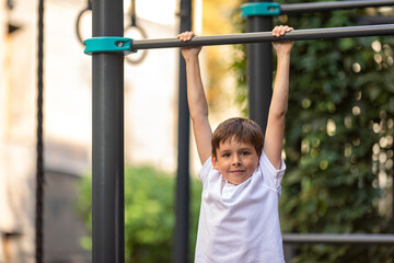 Fototapeta premium Little schoolboy, young athlete is pulling himself up on a sport horizontal bar on outdoor playground on sunny day. Active childhood, acrobat, workout on backyard, summer camp concept