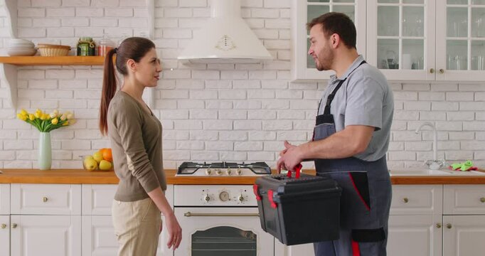 Smiling woman shaking hands with male repairman after repair works in modern kitchen. Professional technician or plumber in uniform holding toolbox, completing home maintenance and assistance service.