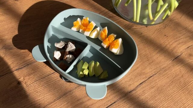 First solid food, healthy baby meal in a silicone plate in the shape of a bear on the table. Beautiful morning sunlight and flowers nearby. 