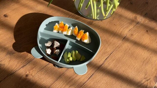 First solid food, healthy baby meal in a silicone plate in the shape of a bear on the table. Beautiful morning sunlight and flowers nearby. 