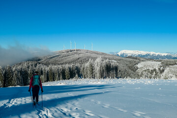 Hiker woman snowshoeing with view of wind turbines stand tall on snow-covered mountain of Fischbach Alps, Styria, Austria. Renewable energy in wilderness. Frosted evergreen forests near Teufelstein