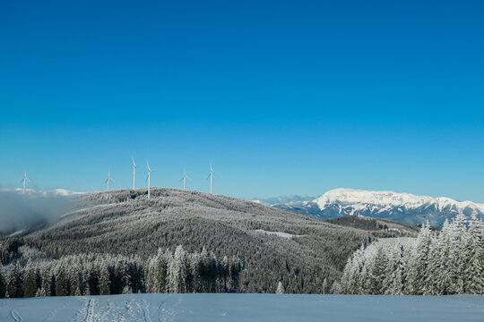 Wind turbines stand tall on snow-covered ridge at Teufelstein, Styria, Austria, harnessing renewable energy amidst serene winter landscape. Frosted trees and distant alpine peaks. Picturesque scene.