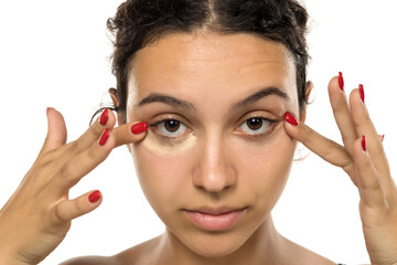 Young woman applying concealer under eyes with fingers. Neutral expression, dark curly hair....