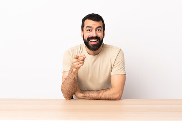 Caucasian man with beard in a table surprised and pointing front.