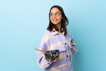 Young brunette mixed race woman holding a bowl full of noodles over isolated blue background with arms crossed and happy.