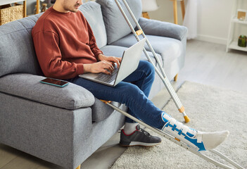 Broken leg or foot injury man working online with laptop, crutches with bandaged leg at home,...