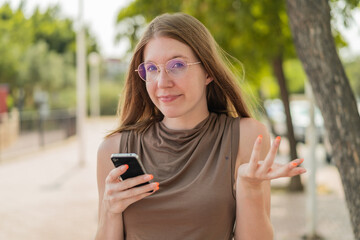 French girl with glasses using mobile phone at outdoors making doubts gesture while lifting the...