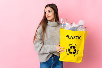 Young caucasian woman holding a bag full of plastic bottles to recycle isolated on pink background...