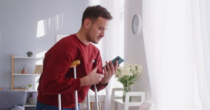 Injured young man posing with crutches, making smartphone call, spending time at home during long recovery, has broken leg after accident. People, health and rehabilitation concept
