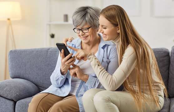 Happy elderly and young women sitting on sofa, using mobile phone together. Adult daughter showing her senior mother how to use smartphone and internet, teaching her to handle with modern device.
