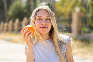 Young blonde woman at outdoors holding an orange
