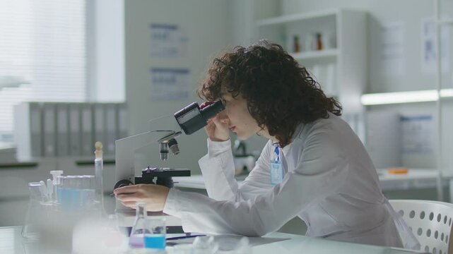 Young female scientist wearing lab coat and badge, analyzing samples under microscope and taking notes at desk with test tubes and flasks on it, doing laboratory research