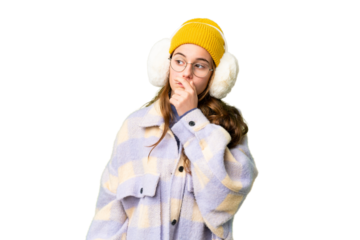 Teenager girl wearing winter muffs over isolated chroma key background having doubts and with confuse face expression