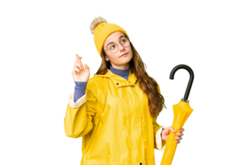 Teenager girl with rainproof coat and umbrella over isolated chroma key background with fingers crossing and wishing the best