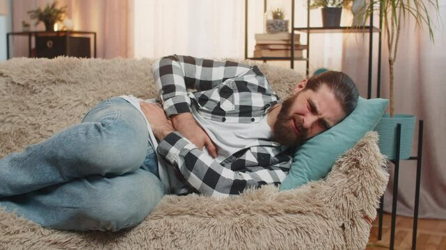 Young ill sick man lying at home with palm on abdomen, quietly moaning from stomach trouble or overeating. Caucasian guy on sofa curls slightly, enduring pain from potential gastritis or diarrhea.