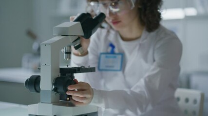 Female scientist wearing protective goggles, adjusting microscope focus during analysis in research laboratory. Shallow focus shot