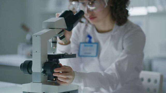 Female scientist wearing protective goggles, adjusting microscope focus during analysis in research laboratory. Shallow focus shot