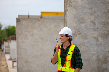 Male civil engineer with walkie talkie and laptop stands next to ready made wall or precast boards conducting a quality check in a factory. Engineer in safety suit working in a precast factory.