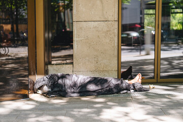 Homeless person resting on sidewalk near modern building while surrounded by urban environment and natural light during the day