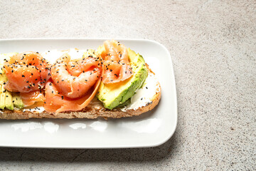 A delicious toast topped with smoked salmon, fresh avocado, cream cheese, and sesame seeds, served on a white plate over a textured neutral background.