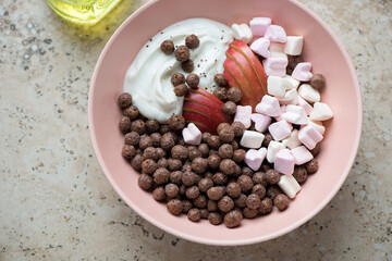 Roseate bowl with chocolate balls, yogurt, marshmallows and apple, horizontal shot on a beige granite surface, high angle view