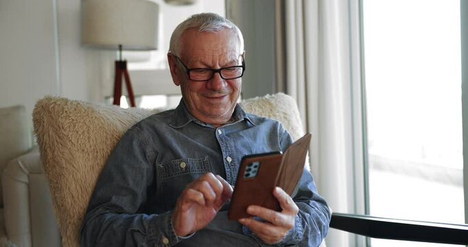 Smiling old retired man wear glasses using smartphone sits in armchair in cozy living room, read and answers on sms, exchanging news with family, enjoy remote communication at home using modern gadget