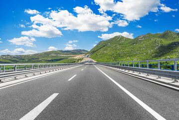 A modern, well-maintained highway stretches into the distance, surrounded by lush green mountains under a bright blue sky with scattered white clouds.
