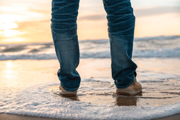 Standing on sandy beach at sunset, person enjoys gentle waves lapping at their feet while the sky is painted in warm hues of orange and pink