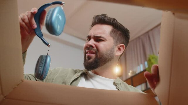 Dissatisfied annoyed male customer open cardboard box receive damaged broken wireless headphones, shocked young man consumer having problem complaint frustrated with bad order delivery at home room.