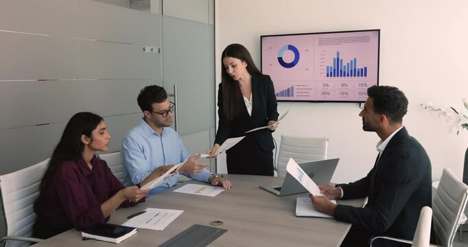 Young Italian business woman distributing printed reports to her colleagues gathered together in boardroom, engaged in discussion, examining documents, and analyzing charts. Training, briefing event