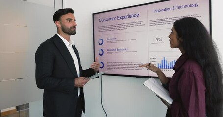 Middle-eastern businessman hold tablet lead talk with Indian businesswoman, present information stand together near large digital screen displayed customer experience, innovation and technology data