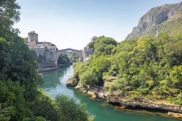 Poster The Stari Most bridge, old bridge on Neretva river in Mostar town, Bosnia and Herzegovina, Europe. Scenic view of a stone bridge and river surrounded by lush greenery, old buildings, and mountains.