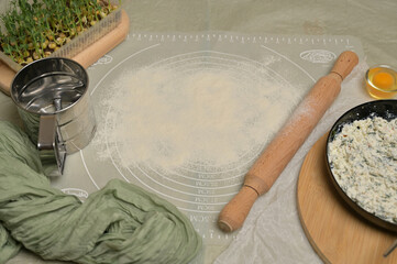 Floured silicone rug on worktop. Rolling pin, sieve, egg, green towel, microgreens sprouts, cottage cheese with parsley and dill greens on kitchen table. Home cooking baking