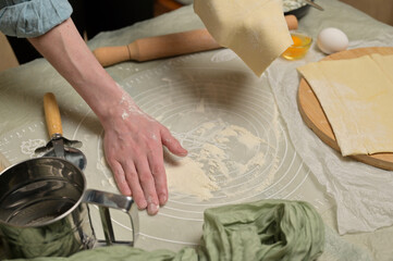 Woman's hand holding a sheet of dough and sprinkles flour worktop. Rolling pin, cutter, eggs, green towel on kitchen table. Home cooking baking puffs