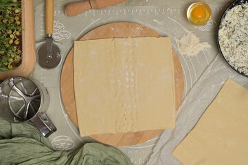 Top view square sheet of dough on wooden board. Rolling pin, cutter, egg, green towel, microgreens sprouts, cottage cheese with parsley and dill greens on kitchen table. Home cooking baking puffs