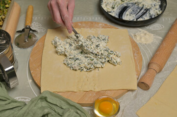 Woman's hand with fork putting cottage cheese with parsley and dill greens on dough. Rolling pin, cutter, egg, green towel and microgreens sprouts on kitchen table. Home cooking baking puffs