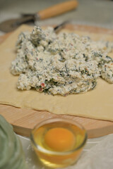 Close up cottage cheese with parsley and dill greens on dough. Wooden board, cutter, egg, green towel on kitchen table. Home cooking baking puffs