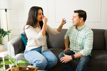 Woman pinching nose, handing breath spray to husband lounging on sofa, indicating unpleasant mouth...