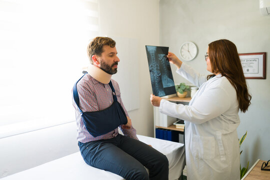 Female doctor showing x-ray scan to male patient wearing neck brace and arm sling in doctor's office during medical consultation