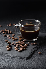 Dark coffee in a glass coffee cup, black espresso and roasted coffee beans pilled on black table,...