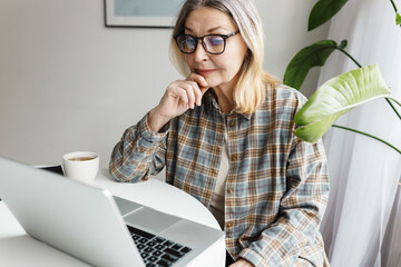 Working pensioner. Senior female CEO or accounting manager using laptop, sitting at table in front of computer, reading documents, or using online application, looking at screen with concentration